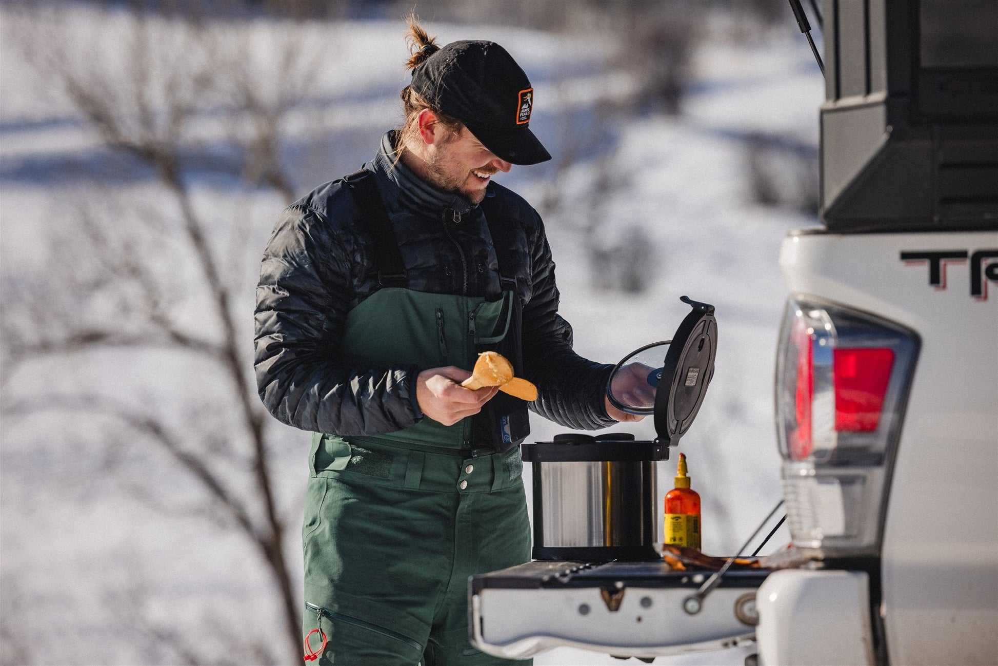 A person in winter clothing uses the RockPot Mini – Off-Grid Cooking, Simplified by RockPot to scoop food from a rice cooker with a spoon on the tailgate of a white truck in a snowy outdoor setting. A sauce bottle sits beside them.