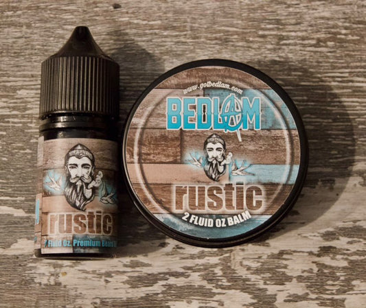 A bottle and tin of Bedlam Beard Companys Rustic beard balm—featuring coffee, tobacco flower, rum, vanilla, and black cherry—sit on wood. Both have labels with a bearded man on rustic wood designs with blue accents.