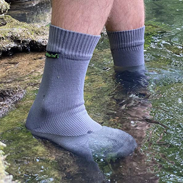 Wearing Pike Trail Waterproof Socks – Stay Dry, Stay Moving, a person stands with both feet in clear water by a rocky, mossy edge outdoors, enjoying comfort and blister protection.