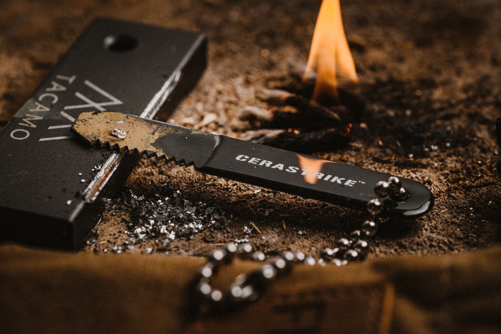 A close-up of the TITAN Survival TACAMO® Mini Fire-Starter Kit with Tinder & Canvas Bag striking a ferro rod on sandy ground, sending sparks near a lit campfire, with a metal bead chain attached to the tool.