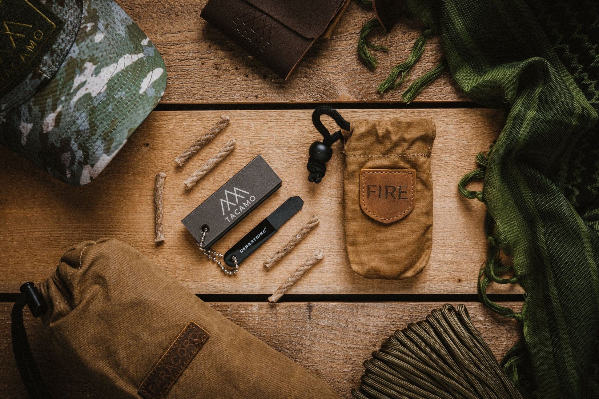 A flat lay of TITAN Survival gear on wood, featuring a camo hat, green scarf, cord, branded tags, drawstring pouches, and the TACAMO® Mini Fire-Starter Kit with Tinder & Canvas Bag.