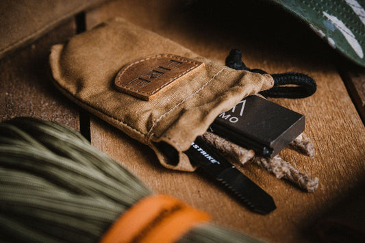 The TITAN Survival TACAMO® Mini Fire-Starter Kit with Tinder & Canvas Bag includes a black ferro rod and striker, shown on wood next to green paracord and a camouflage item.