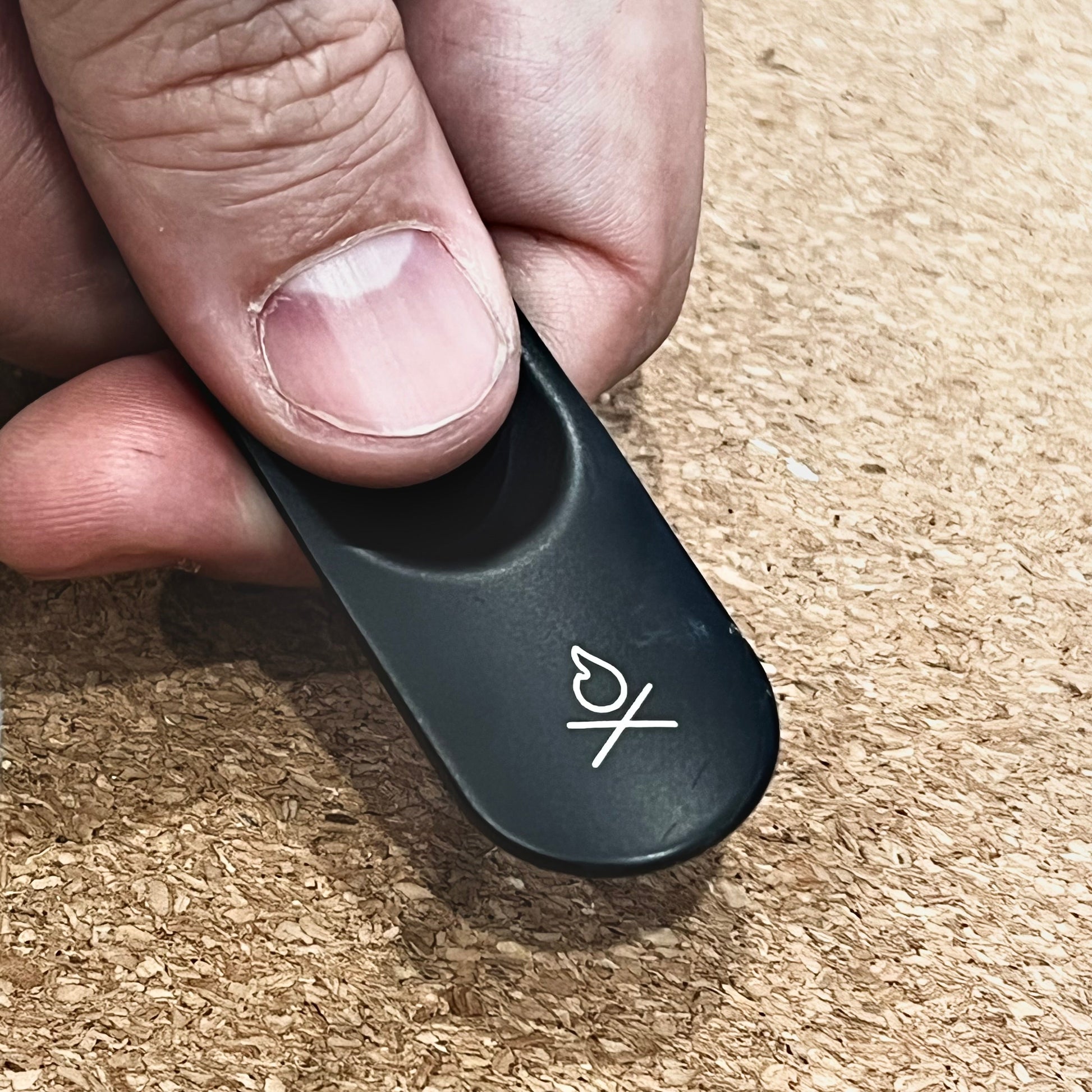 A close-up of a hand holding the Überleben Zünden PRO Modern Flint & Steel Fire Starter by Überleben, featuring a white heart and “X” symbol, against a textured brown cork background.