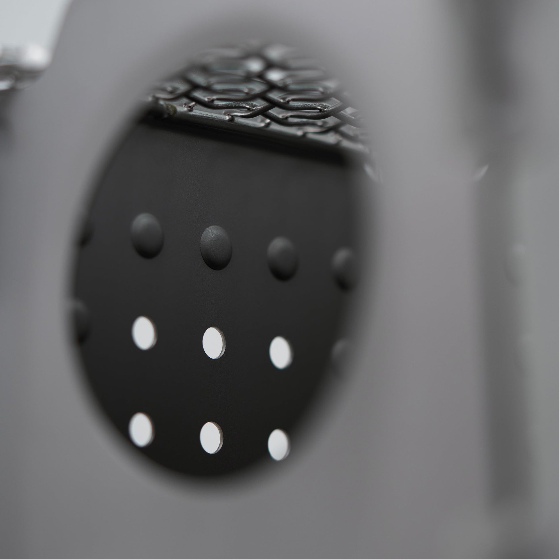A close-up of the Überleben Stöker Flatpack Stove by Überleben shows its circular metal surface with evenly spaced holes, partially obscured in the foreground. The out-of-focus background displays a subtle grid-like pattern.