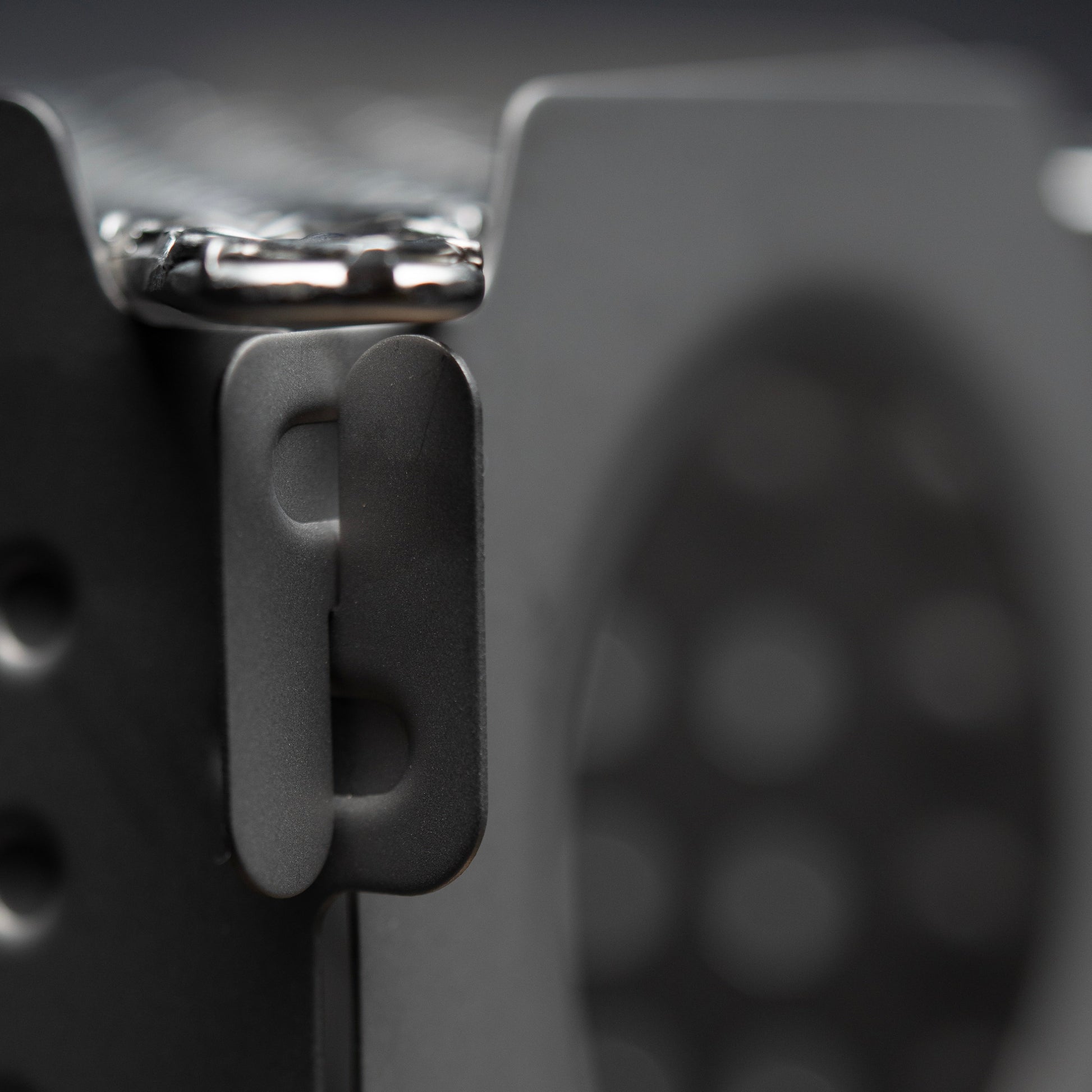 Close-up of a matte black metal latch on the Überleben Stöker Flatpack Stove by Überleben, shown attached to its perforated surface with a blurred patterned background.