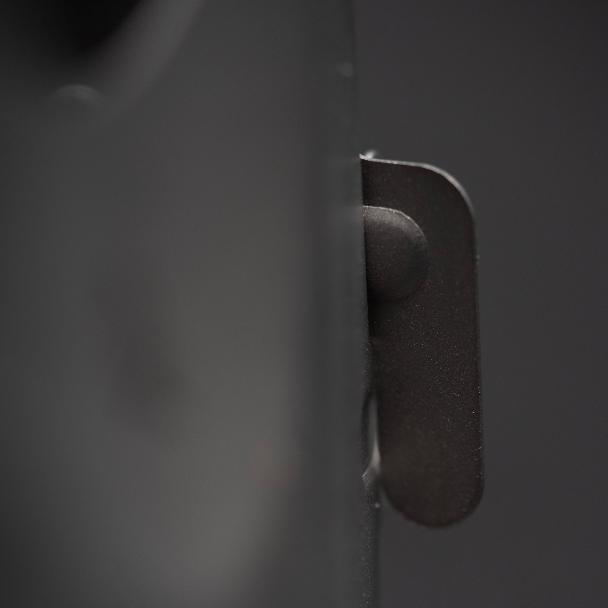 Close-up of a dark metal latch protruding from the smooth surface of the Überleben Stöker Flatpack Stove by Überleben, with a blurred, dark background.
