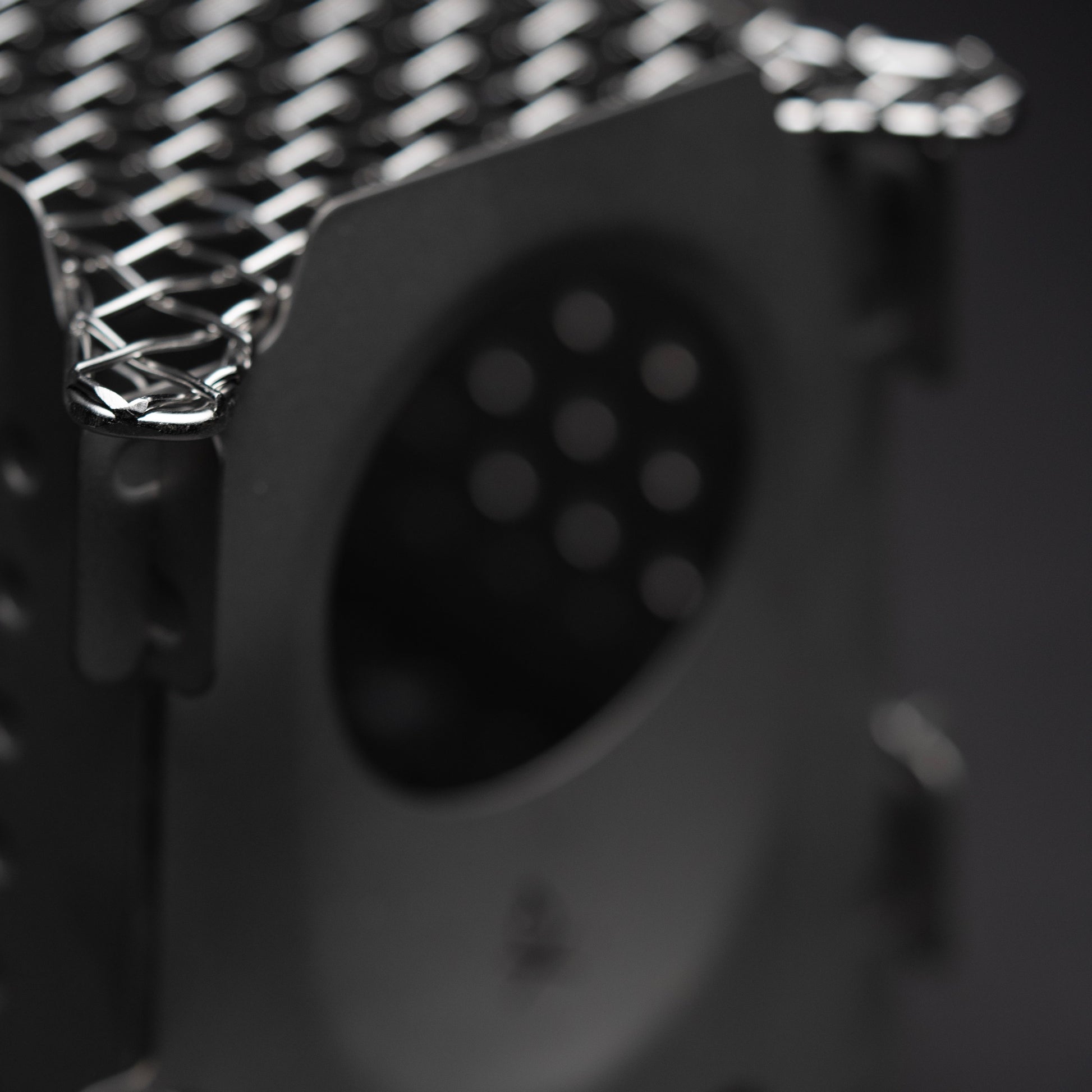 Close-up of the Überleben Stöker Flatpack Stove by Überleben, featuring a metallic, perforated grid top and side opening. The image highlights this survival tools textured finish and geometric design against a dark, blurred background.