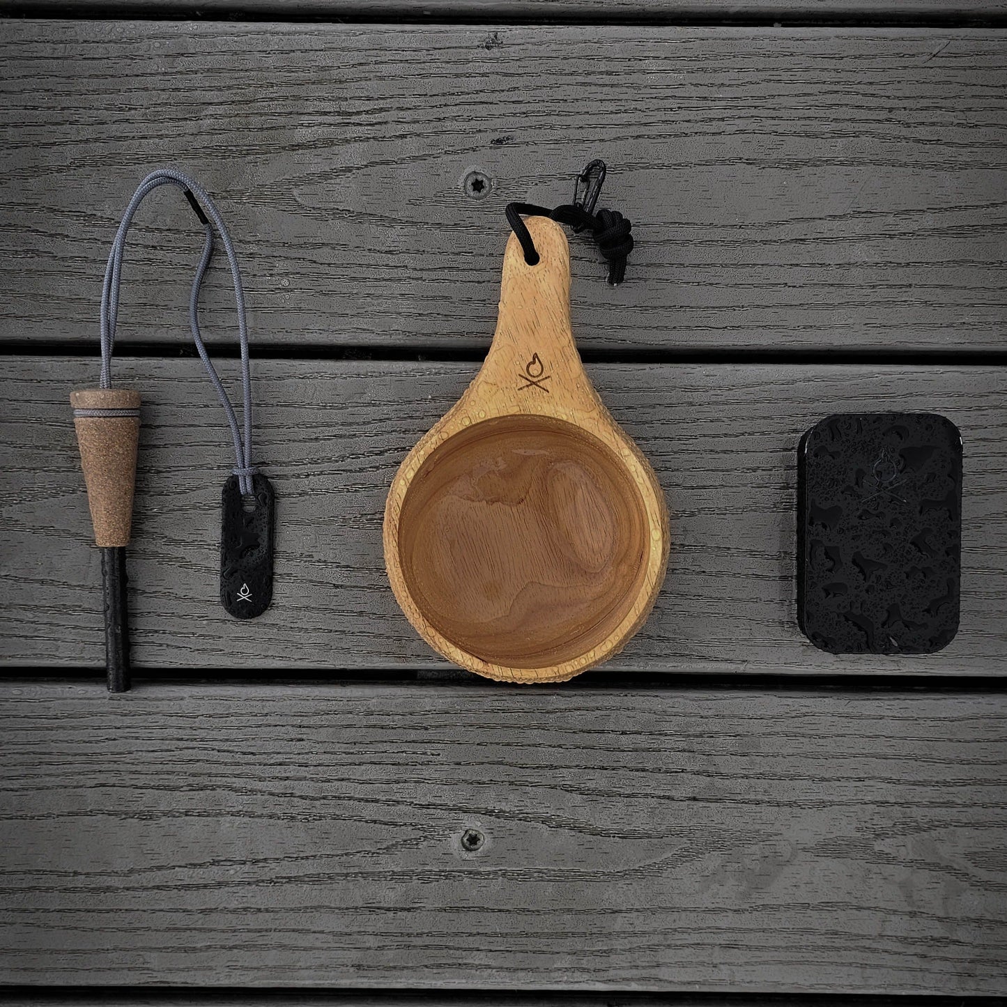Überleben’s The Lore – Oversized Hardwood Kuksa, a fire starter with cork handle and lanyard, and a black tin are neatly arranged on a gray wooden surface.