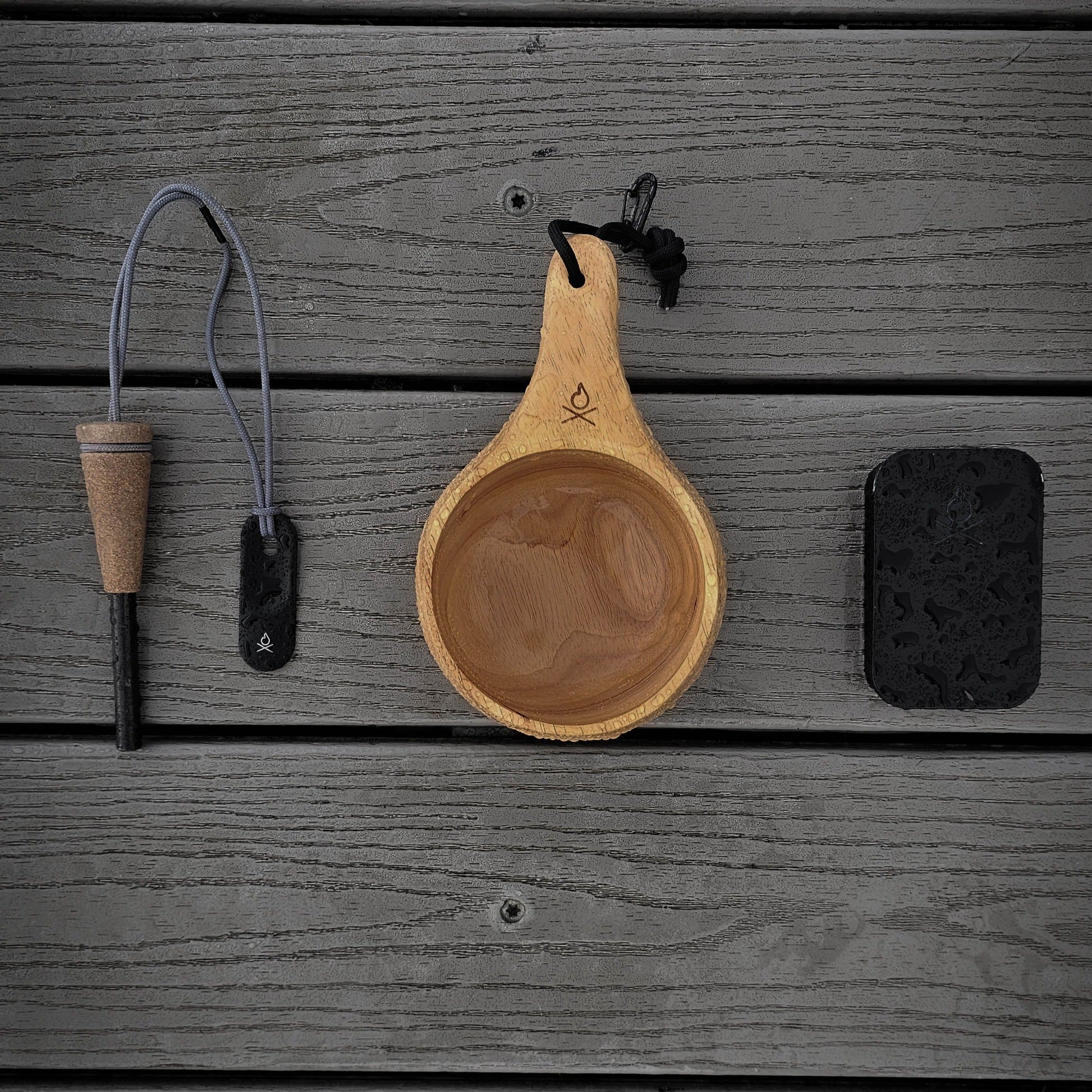 Überleben’s The Lore – Oversized Hardwood Kuksa, a fire starter with cork handle and lanyard, and a black tin are neatly arranged on a gray wooden surface.