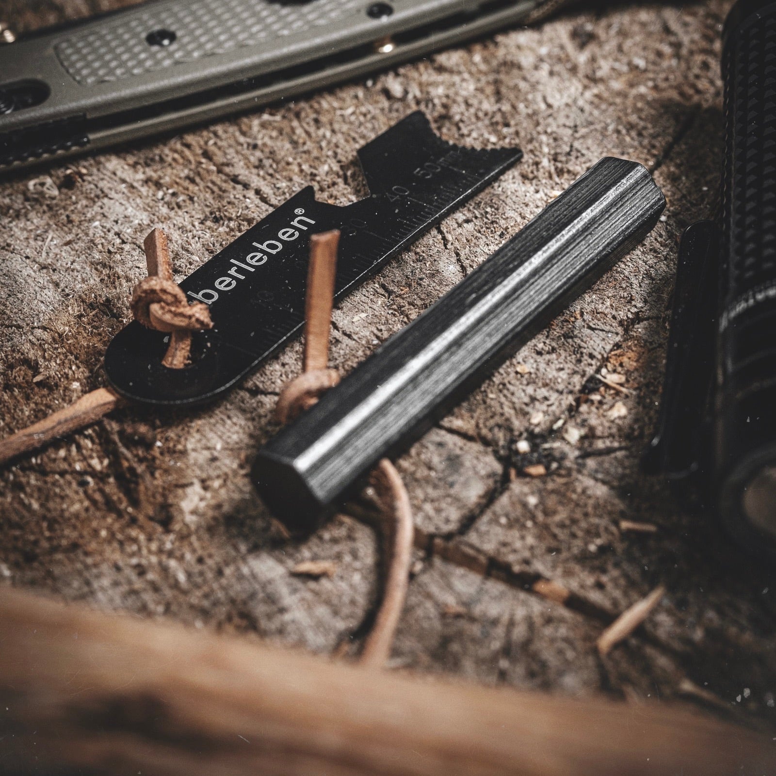 A close-up of survival tools on wood: the Überleben Hexå Original Fire Starter, a metal striker labeled “überleben,” and a flashlight, all tied with leather cords.