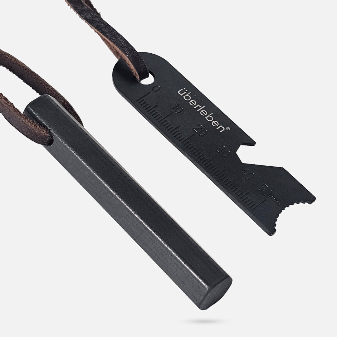 The Überleben Hexå Original Fire Starter features a black ferrocerium rod and a black multi-tool scraper with ruler markings and “überleben” branding, both attached to a brown leather cord.