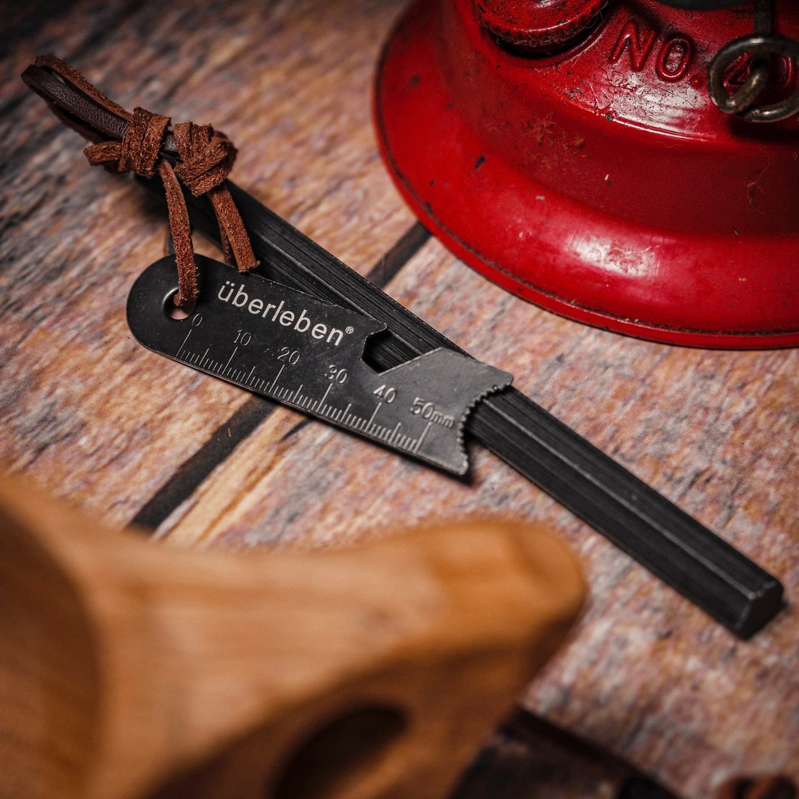 A black Überleben Hexå Original fire starter with a ruler printed on it, attached to a leather cord, rests on a wooden surface beside a red metal item.