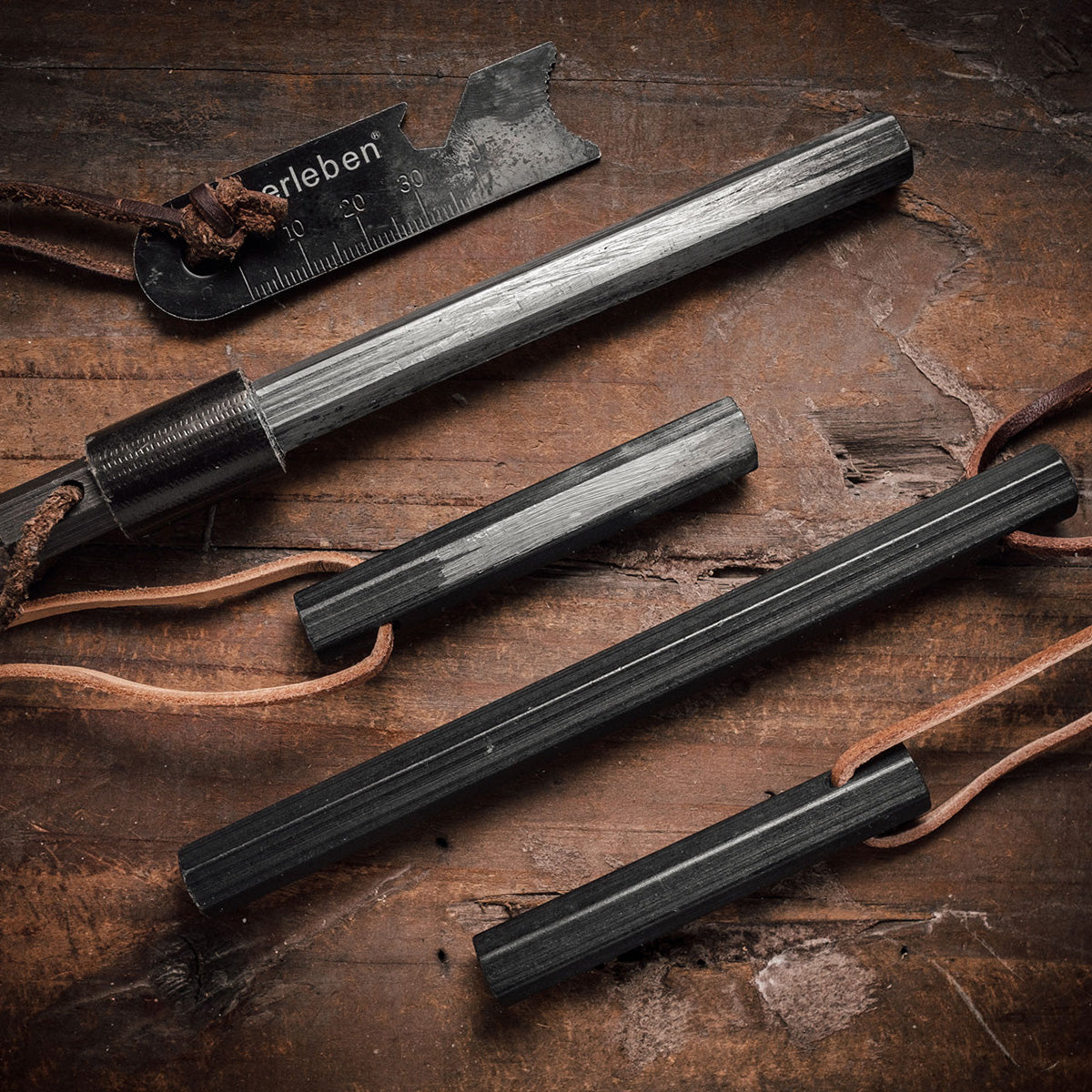 The Überleben Hexå Original Fire Starter set includes four black ferrocerium rods with leather lanyards and a black multi-tool scraper striker with measurement markings, all displayed on a rustic wooden surface.