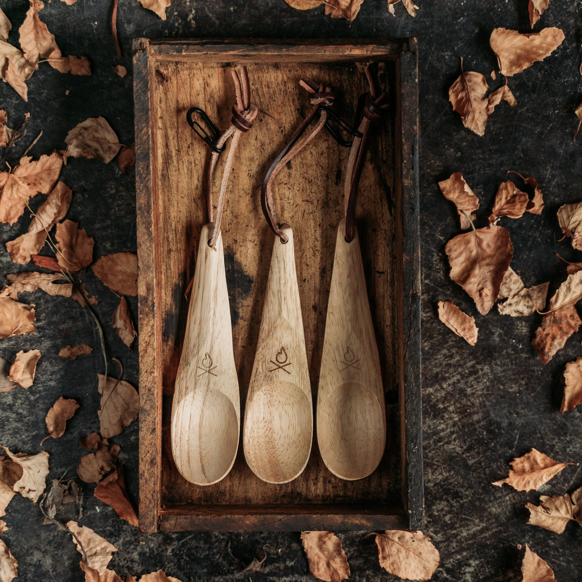 Three Überleben Kanu Hardwood Camping Spoons by Überleben, each with a leather strap, are arranged in a rustic wooden tray and surrounded by dry brown leaves on a dark background.