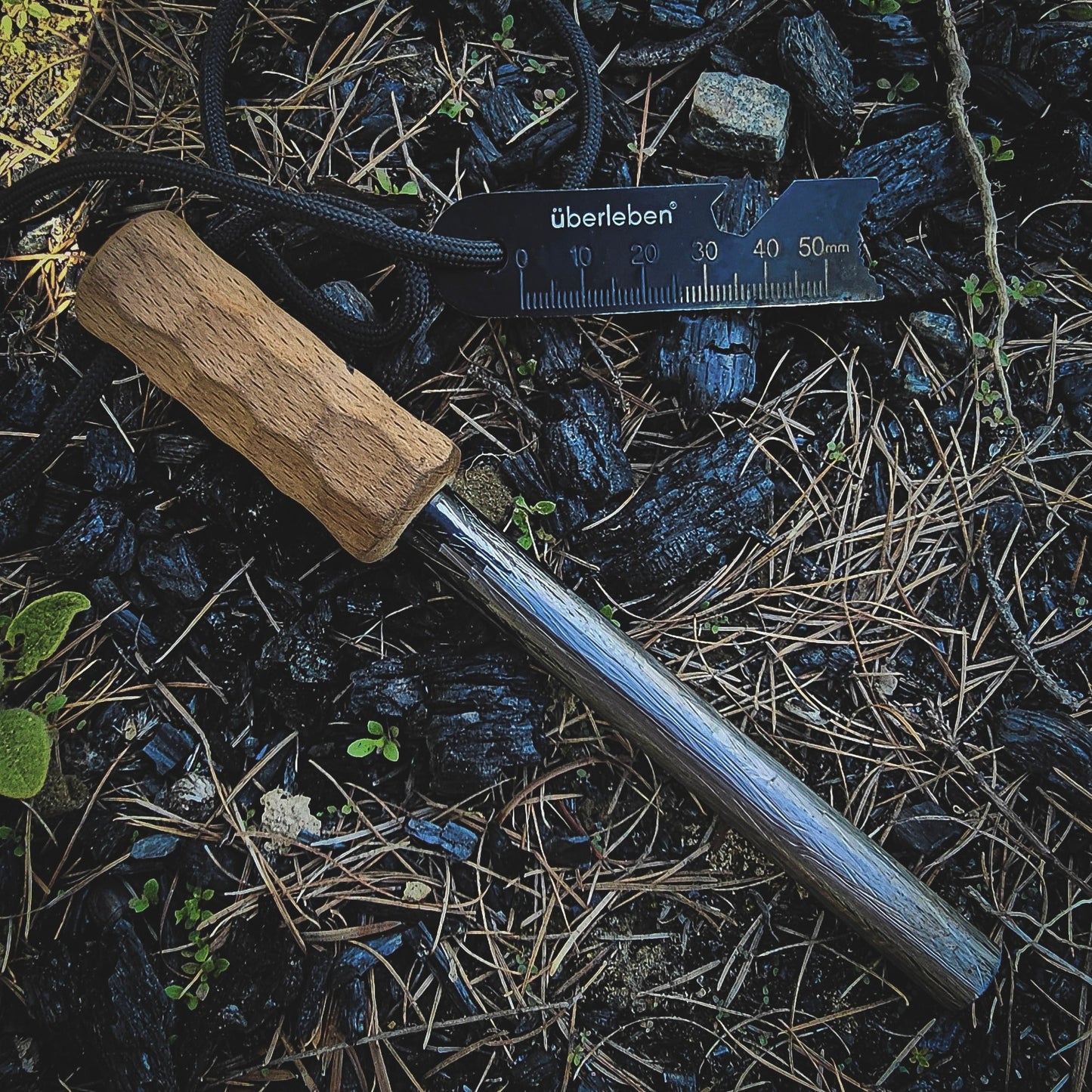The Überleben Kräftig Grip (PRO) – The Heavy-Duty Ferro Beast, featuring a wooden handle and multi-tool striker, rests on charred ground beside a black Überleben ruler, surrounded by pine needles and green plants.
