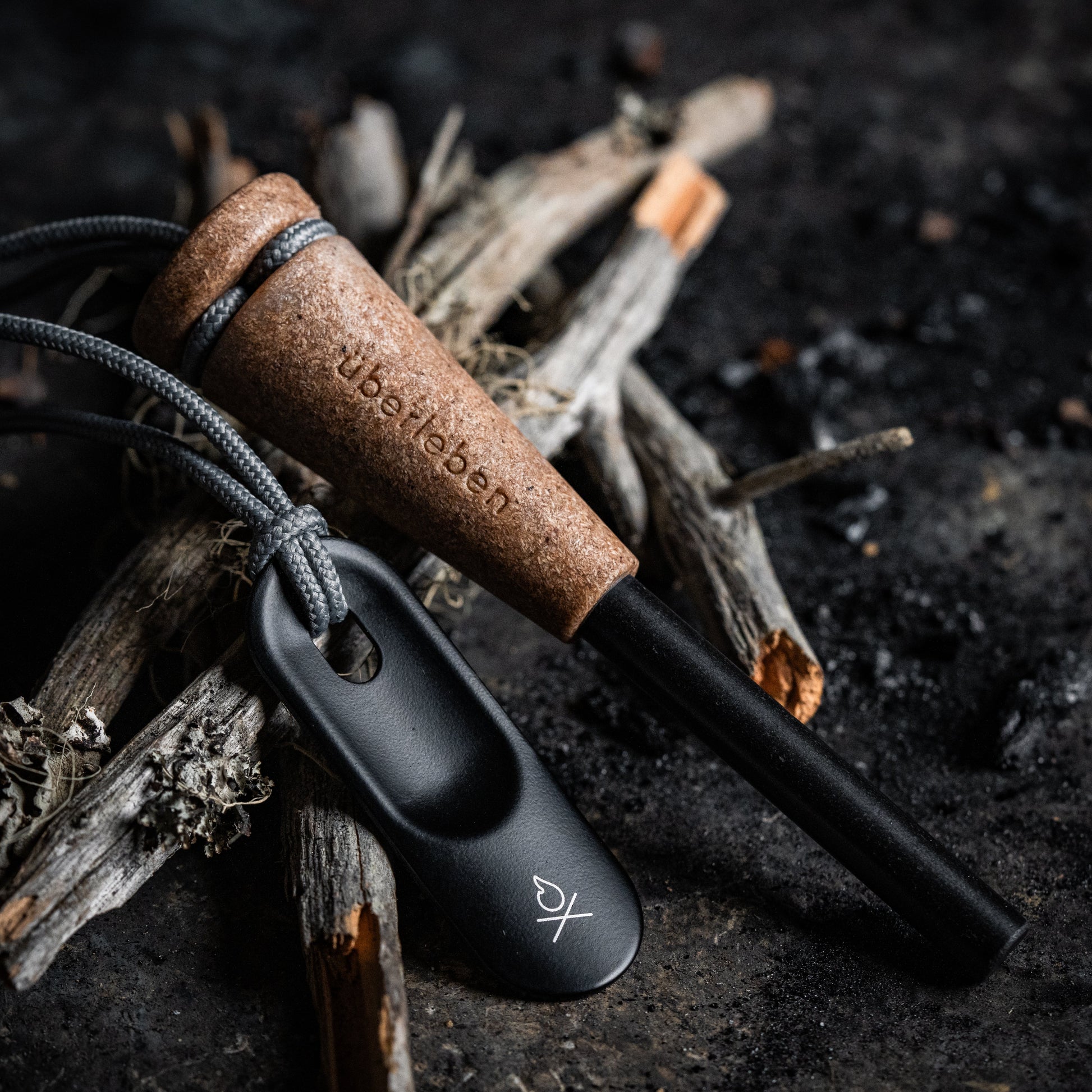 The Überleben Zünden PRO Modern Flint & Steel Fire Starter, with a brown wood-composite handle engraved with the Überleben logo, plus a gray cord and black striker, rests on dry sticks and charcoal on a dark surface.