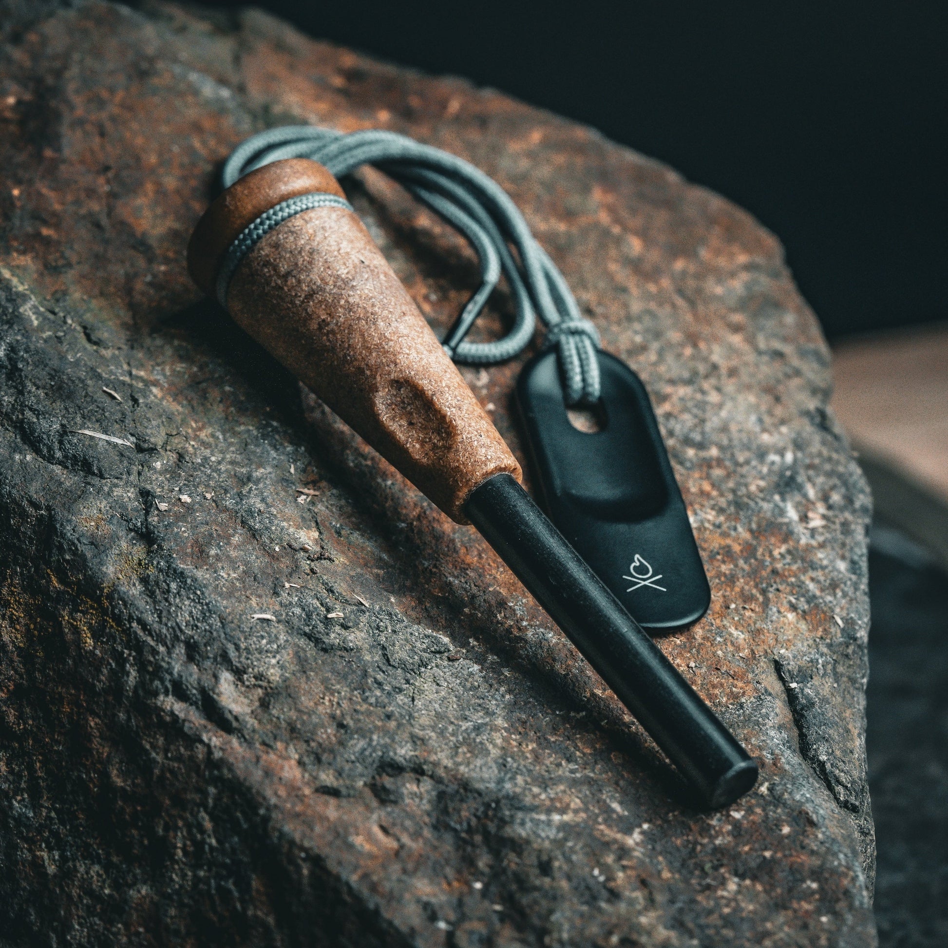 The Überleben Zünden PRO Modern Flint & Steel Fire Starter, featuring a wooden handle and tied cord, rests on a large textured rock with its black striker (with a small logo, made in USA) attached beside it.