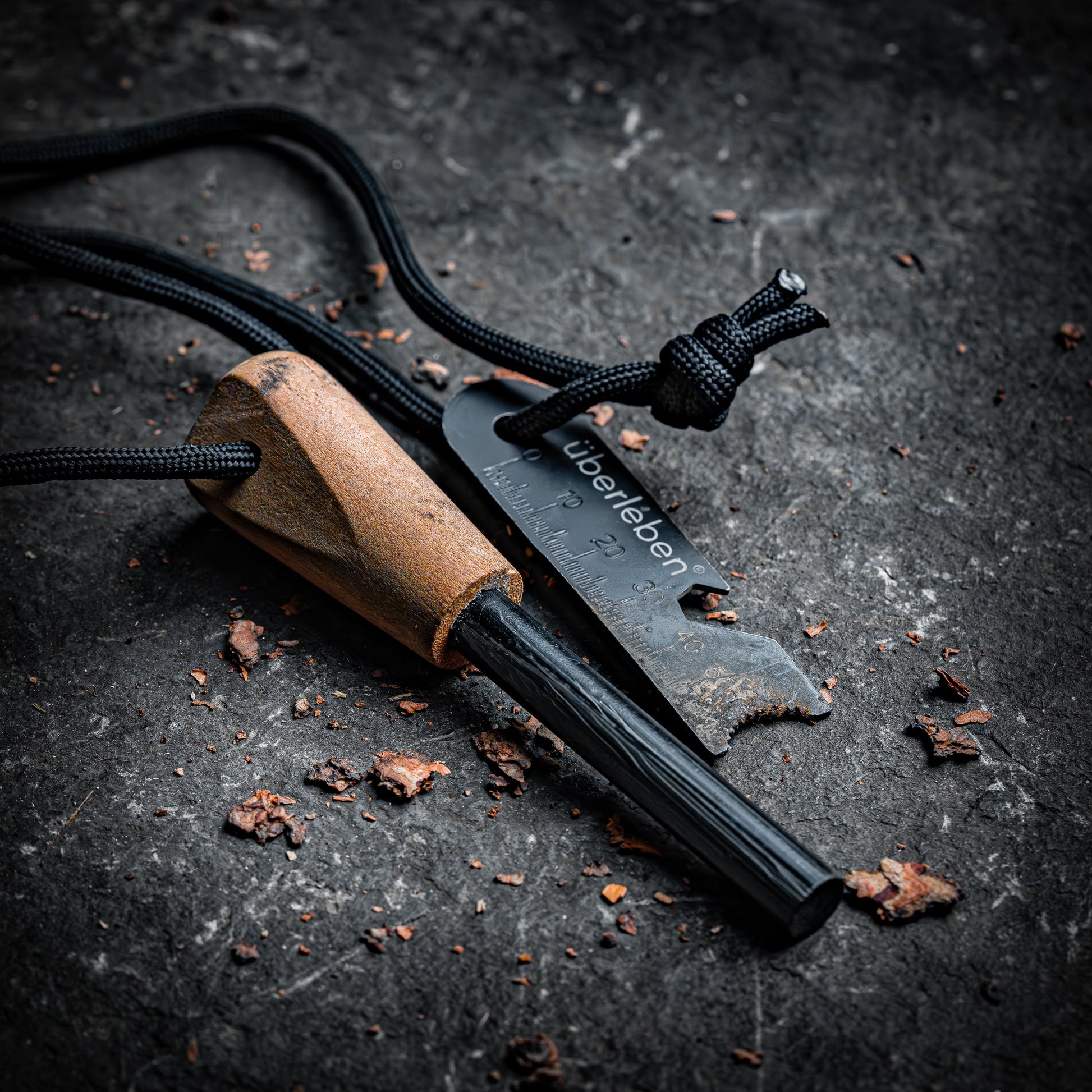The Überleben Zünden Original – Bushcraft Fire Starter by Überleben features a ferro rod with a wooden handle and black striker, both on a black cord, set on a rugged surface—an essential survival tool for any adventure.