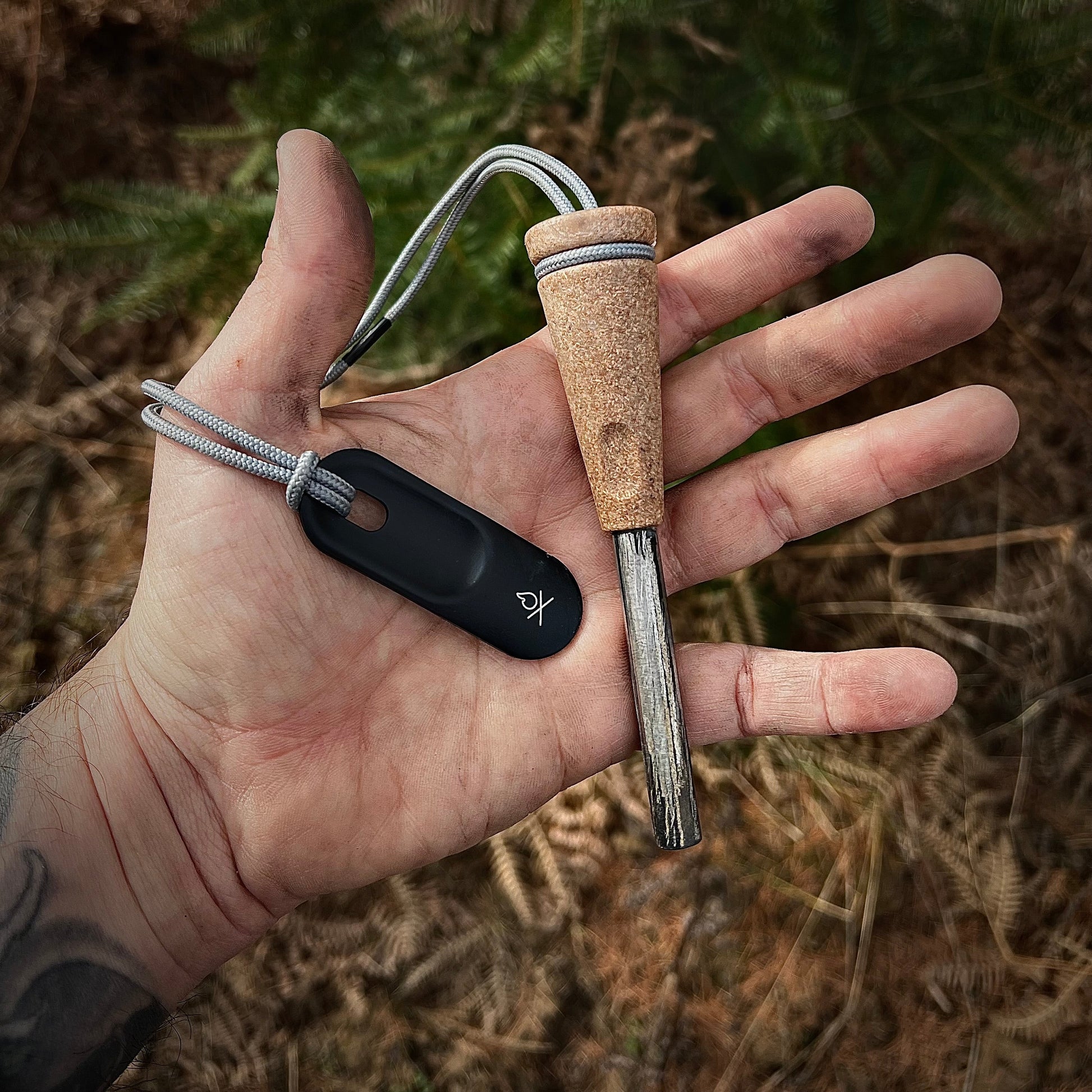 A hand holds the Überleben Zünden PRO Modern Flint & Steel Fire Starter by Überleben, featuring a wooden handle, metal rod, black striker on a gray cord. Ferns and plants blur in the background. Made in the USA for reliable fire starting.