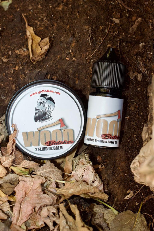 A tin of Bedlam Beard Company’s Wood Butcher beard balm and a bottle of conditioning oil, featuring an axe and bearded man logo, sit on soil with dry leaves. Scent notes: mahogany, teakwood, vanilla, oak, amber, and musk.